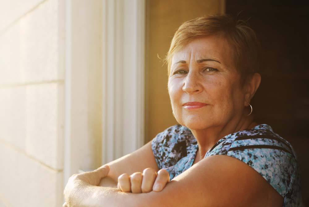 Senior leans in window frame, bathed in warm sunlight.