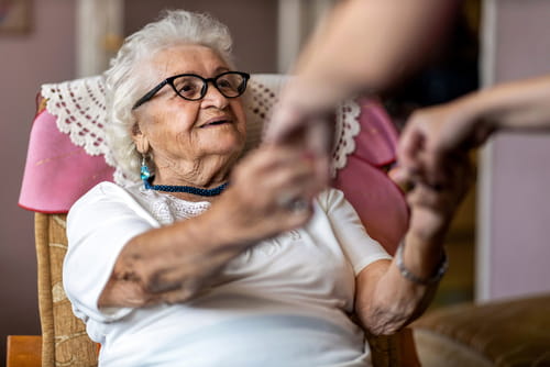 Home caregiver assists senior woman in standing up from armchair.